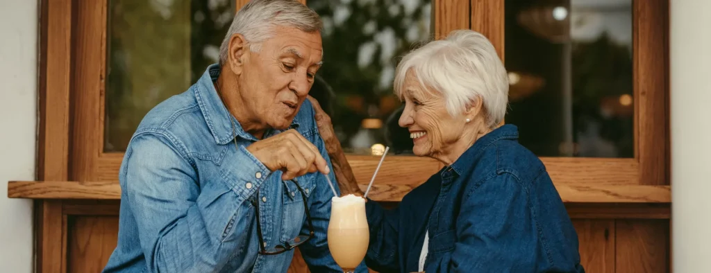 Elderly couple in denim jackets enjoying a creamy drink with straws at a cafe with wooden window background.