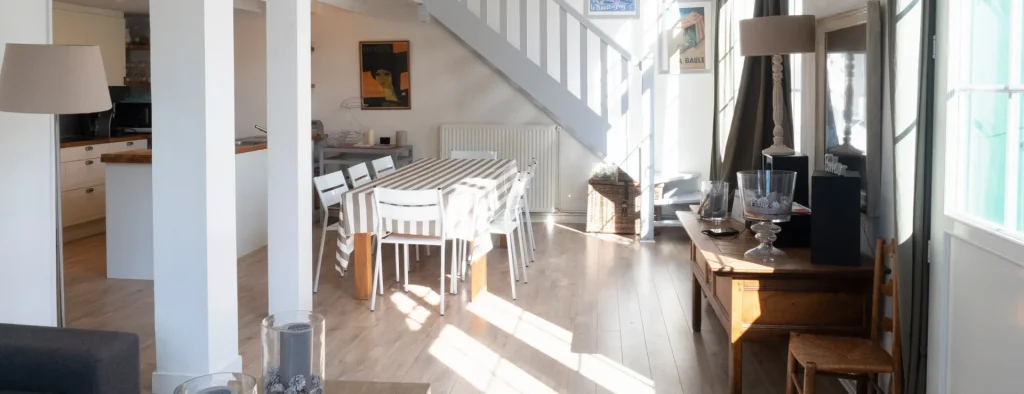Bright dining area with striped tablecloth, white chairs, wooden floor, staircase, and sunlit windows.